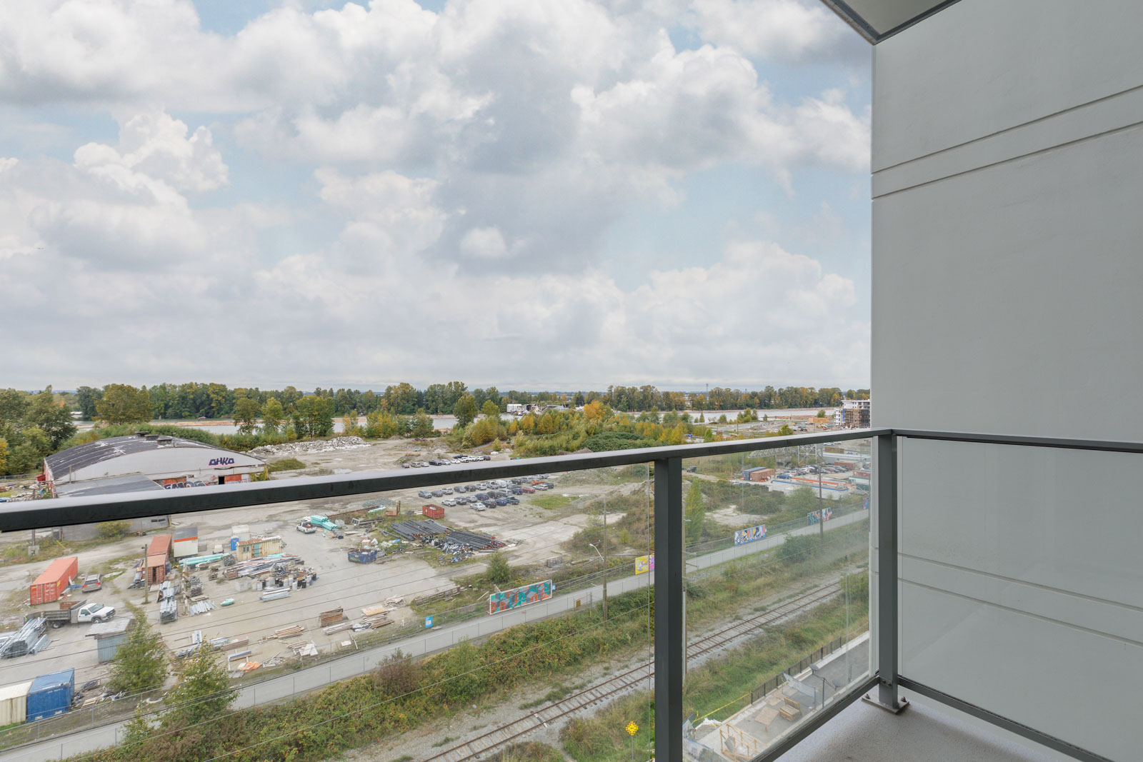View from a balcony showing a construction site with scattered containers and equipment, railroad tracks, and a tree line under a cloudy sky.