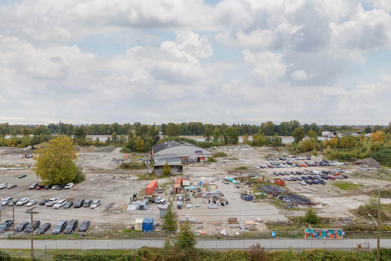 Aerial view of a large construction site with scattered vehicles, equipment, and materials. Trees and a river appear in the background under a cloudy sky.