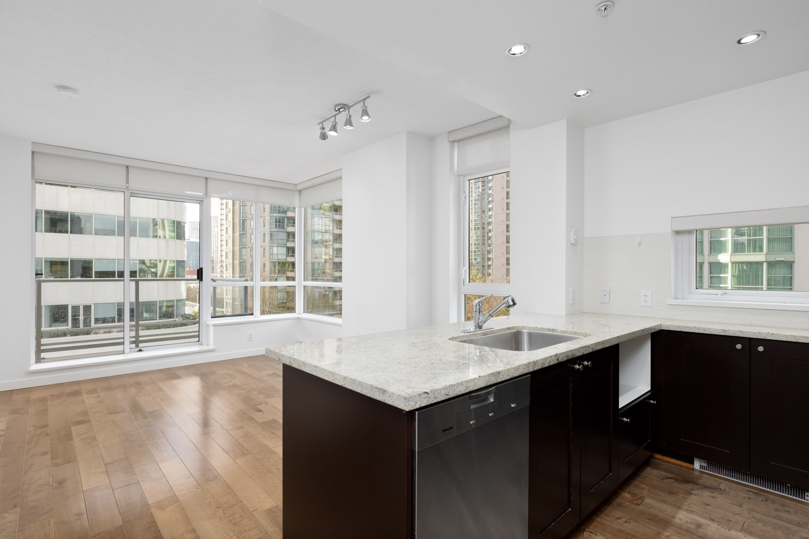 Modern apartment interior with hardwood floors, large windows, and an open kitchen featuring dark cabinets and a granite countertop with a sink.