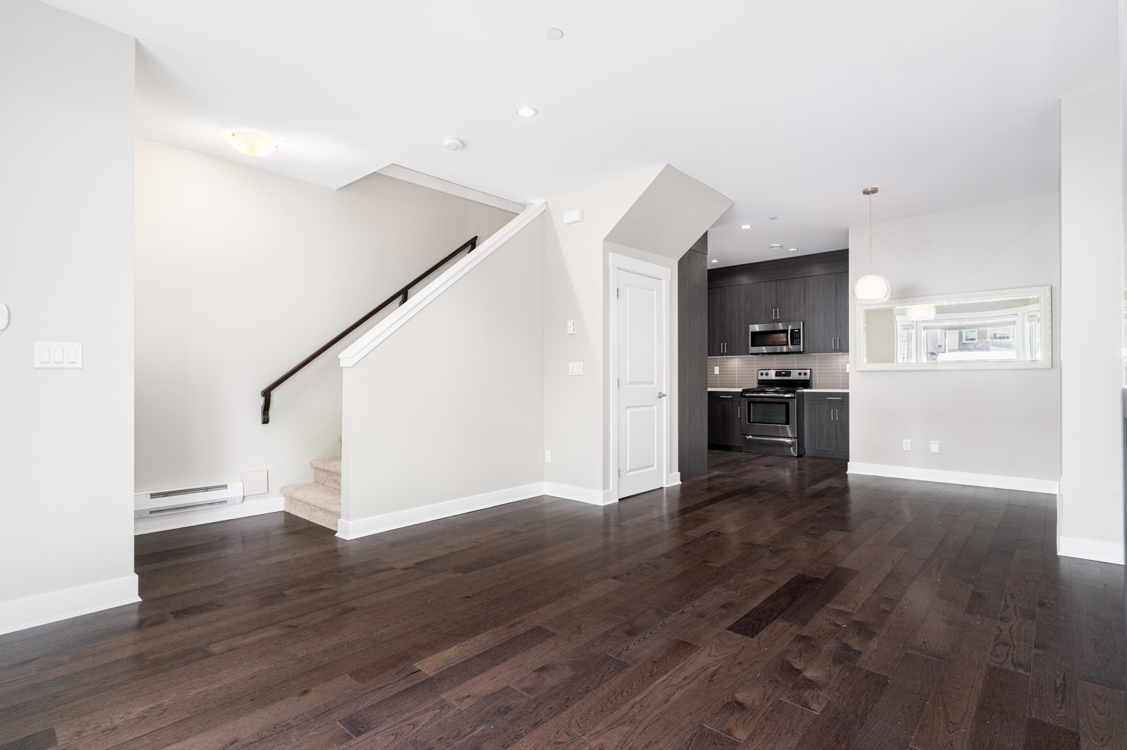A modern, empty room with dark hardwood floors, white walls, a staircase, and a kitchen with dark cabinets and stainless steel appliances.