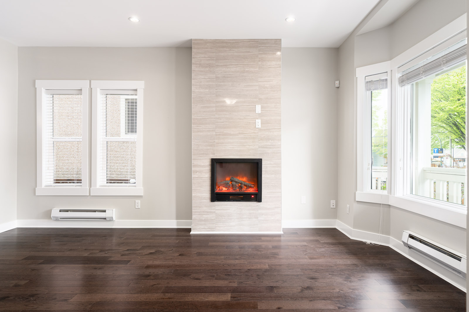 A modern living room with a sleek electric fireplace, large windows with blinds, and dark wooden floors.