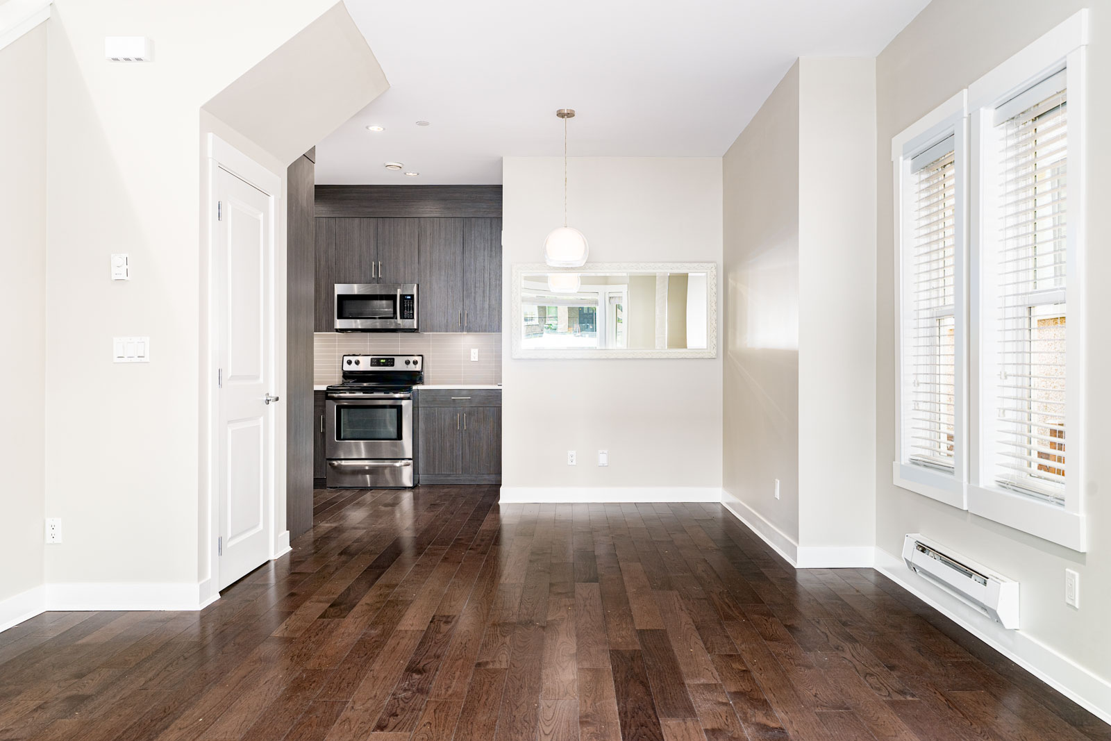 Empty modern kitchen and dining area with dark wood flooring, white walls, stainless steel appliances, a hanging light fixture, large window, and a wall mirror.