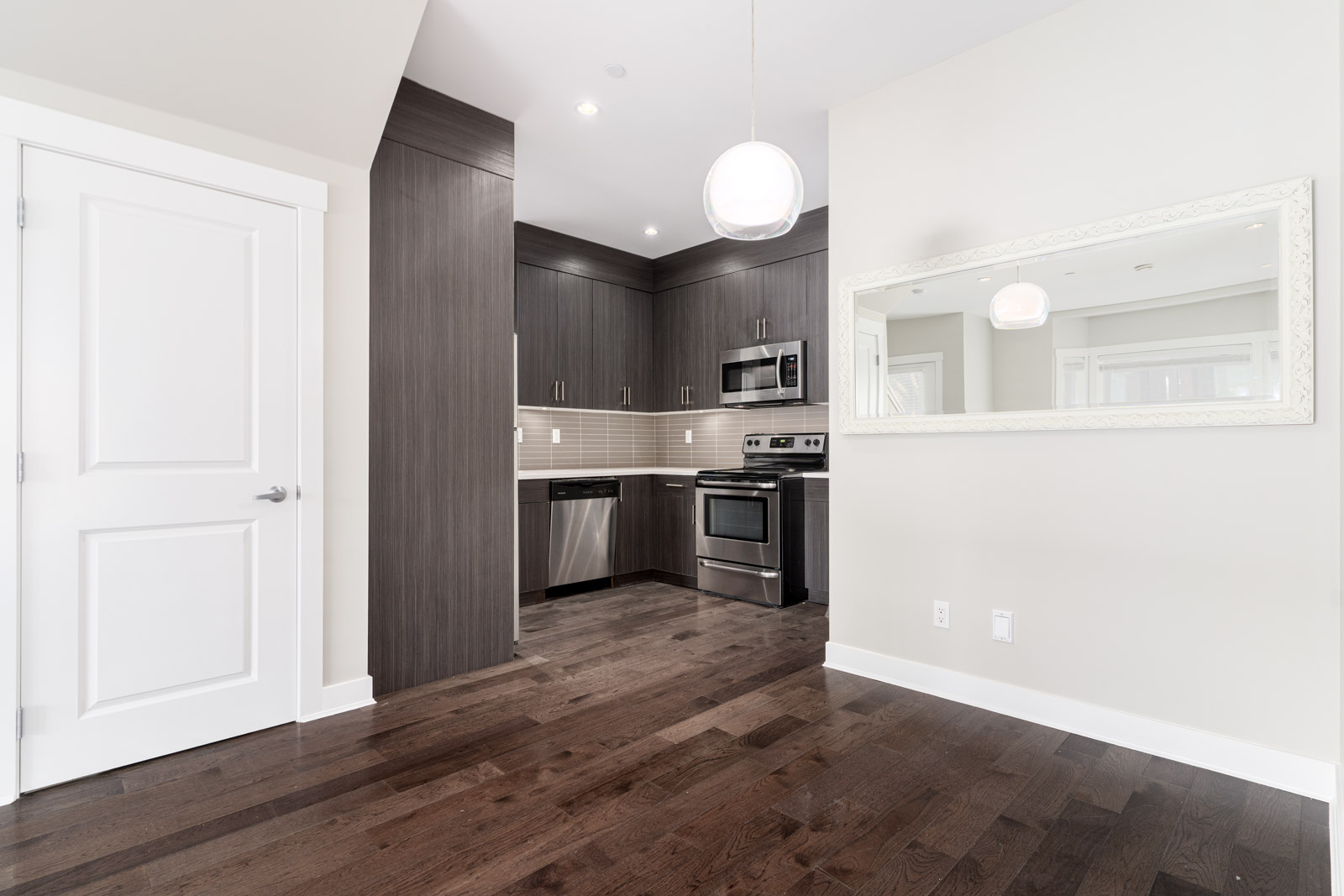 A modern kitchen with dark wood cabinets, stainless steel appliances, and a white-framed mirror on the wall. The floor is dark hardwood, and a white door is on the left.