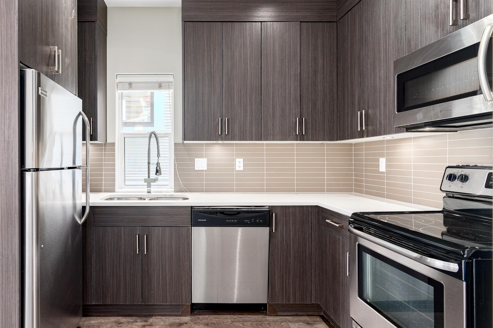 Modern kitchen with dark wood cabinets, stainless steel appliances, and a tiled backsplash. Includes a sink, dishwasher, fridge, oven, and microwave.