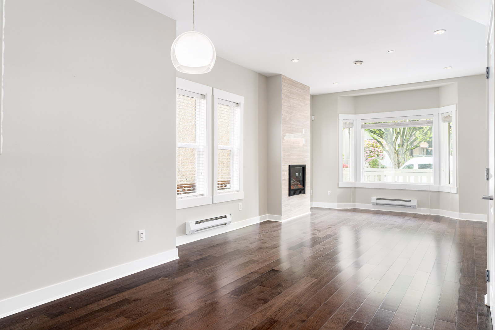 Bright, empty living room with dark hardwood floors, white walls, a fireplace, two large windows, and a hanging light fixture.
