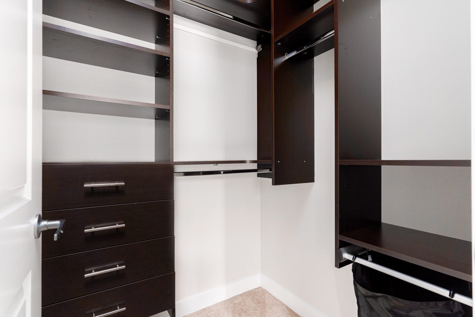 Empty walk-in closet with dark wood shelves, drawers, and hanging rods. White walls and beige carpet.