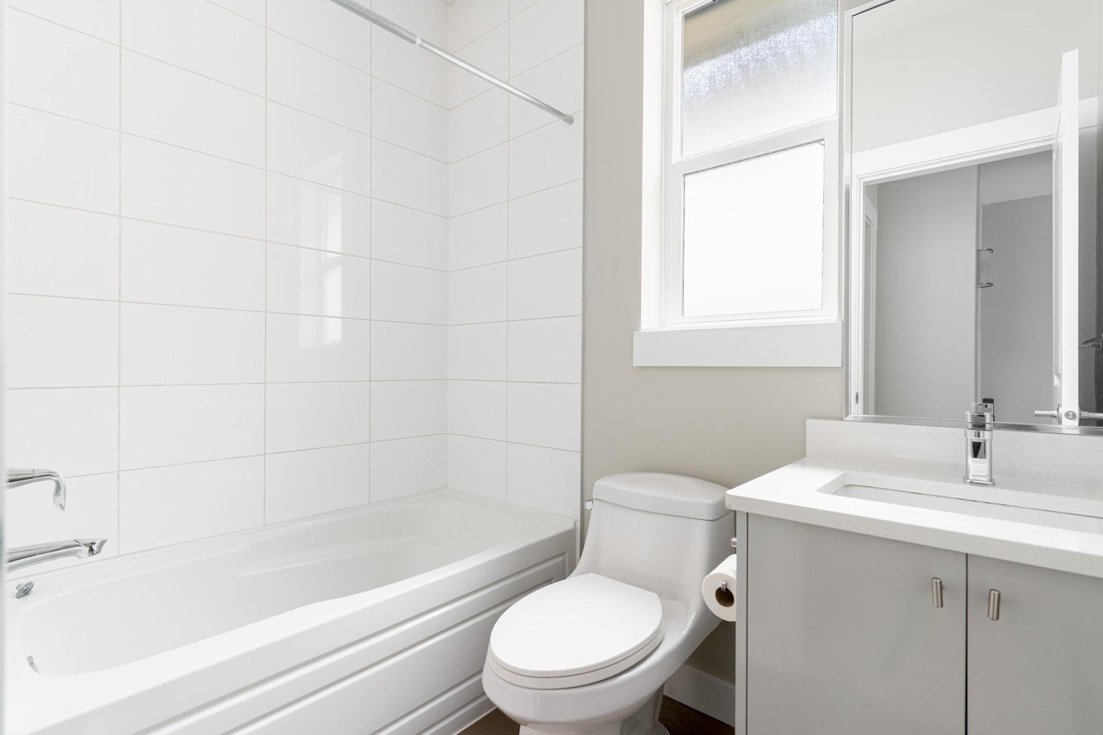 Modern bathroom with white tiles, featuring a bathtub, toilet, and sink with a mirror. A window allows natural light.