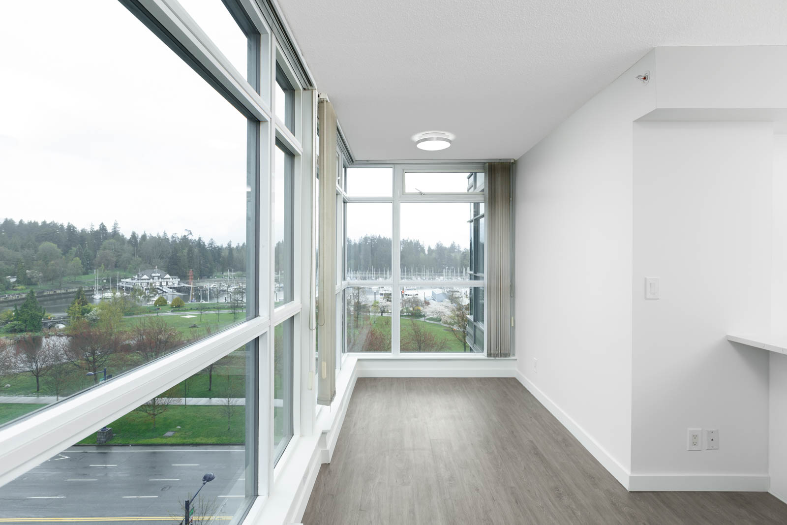 A bright, empty room with large floor-to-ceiling windows, light gray walls, and wood flooring, overlooking a park and marina outside.