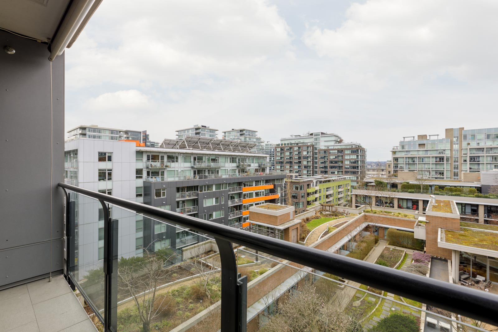 View from a balcony overlooking modern apartment buildings with glass facades and green rooftops under a cloudy sky.