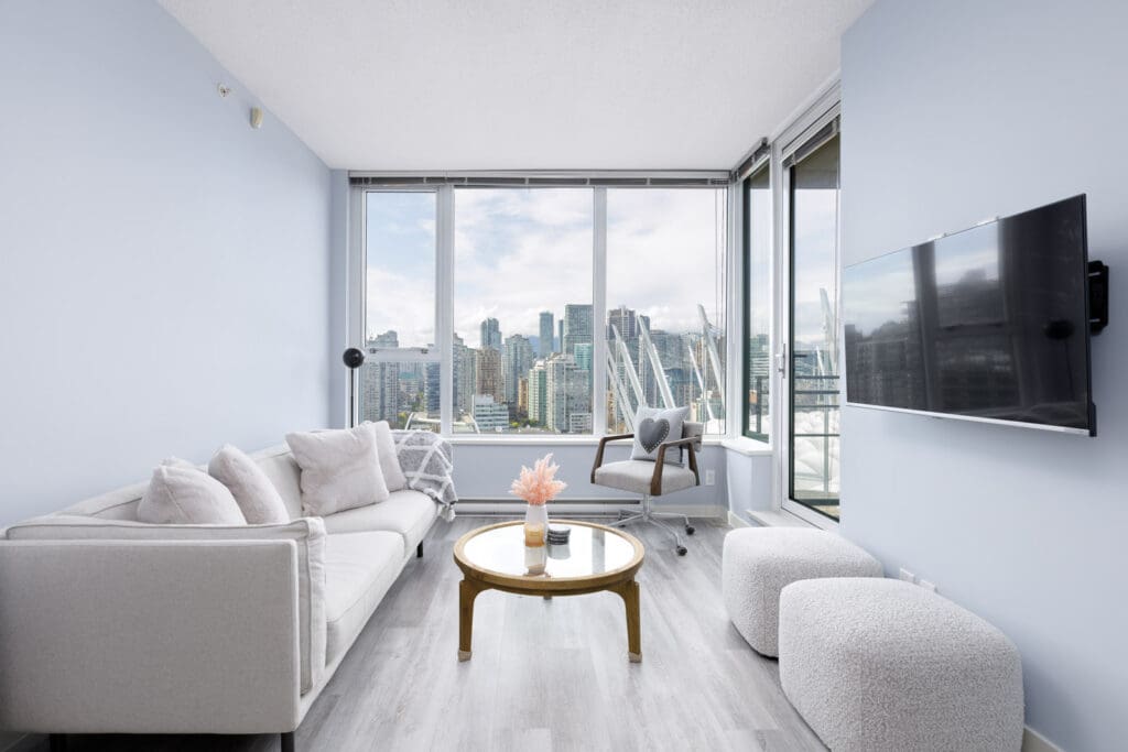 Modern living room with light gray sofas, a small round coffee table, and a wall-mounted TV. Large windows offer a city skyline view. Walls are light blue, and the floor is wood.