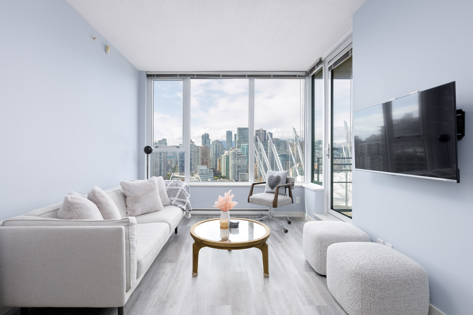 Modern living room with light gray sofas, a small round coffee table, and a wall-mounted TV. Large windows offer a city skyline view. Walls are light blue, and the floor is wood.