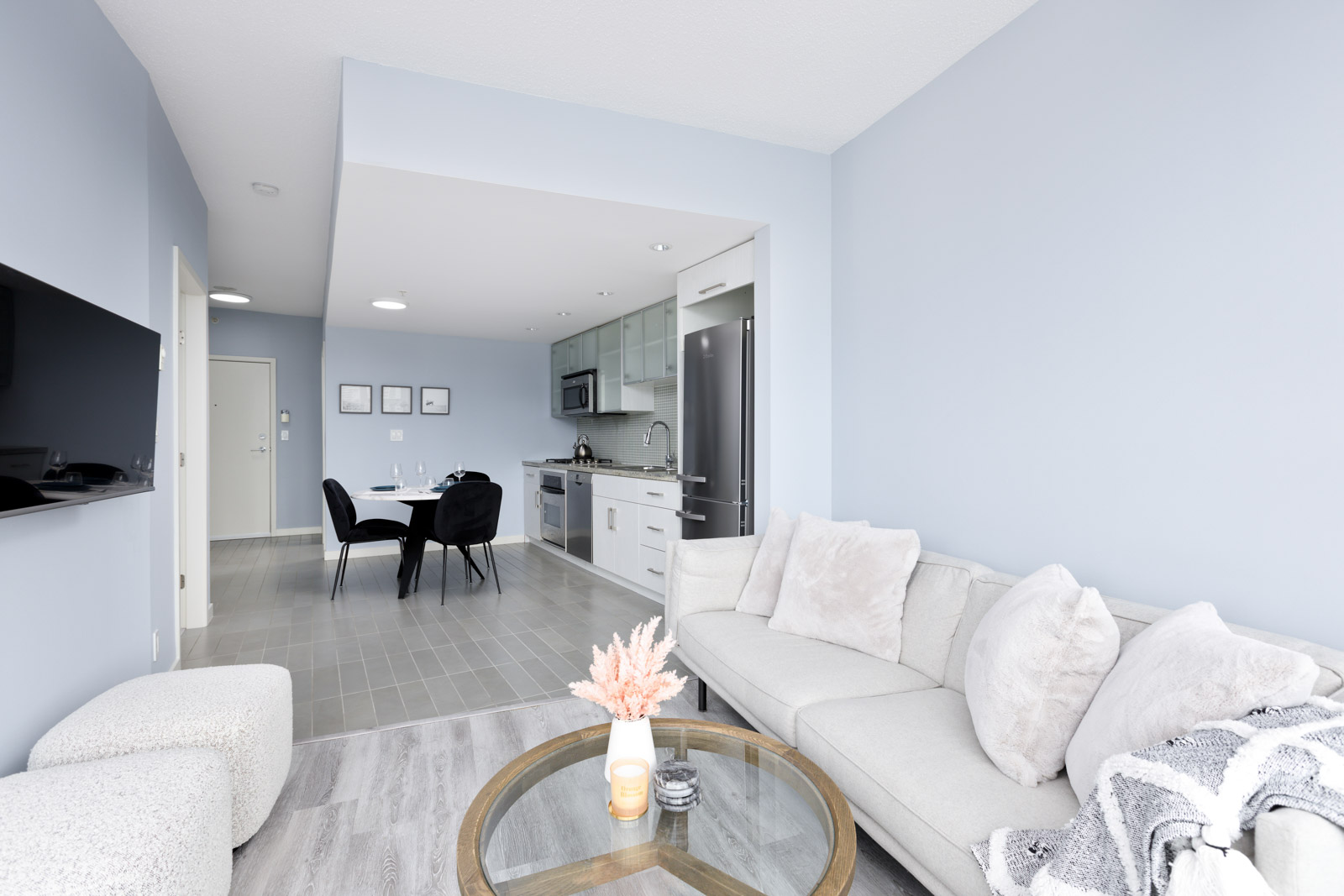 Modern open-plan living space with light blue walls. Features a beige sofa, round glass coffee table, and TV. Dining area with black chairs and table. Kitchen in background with white cabinets.
