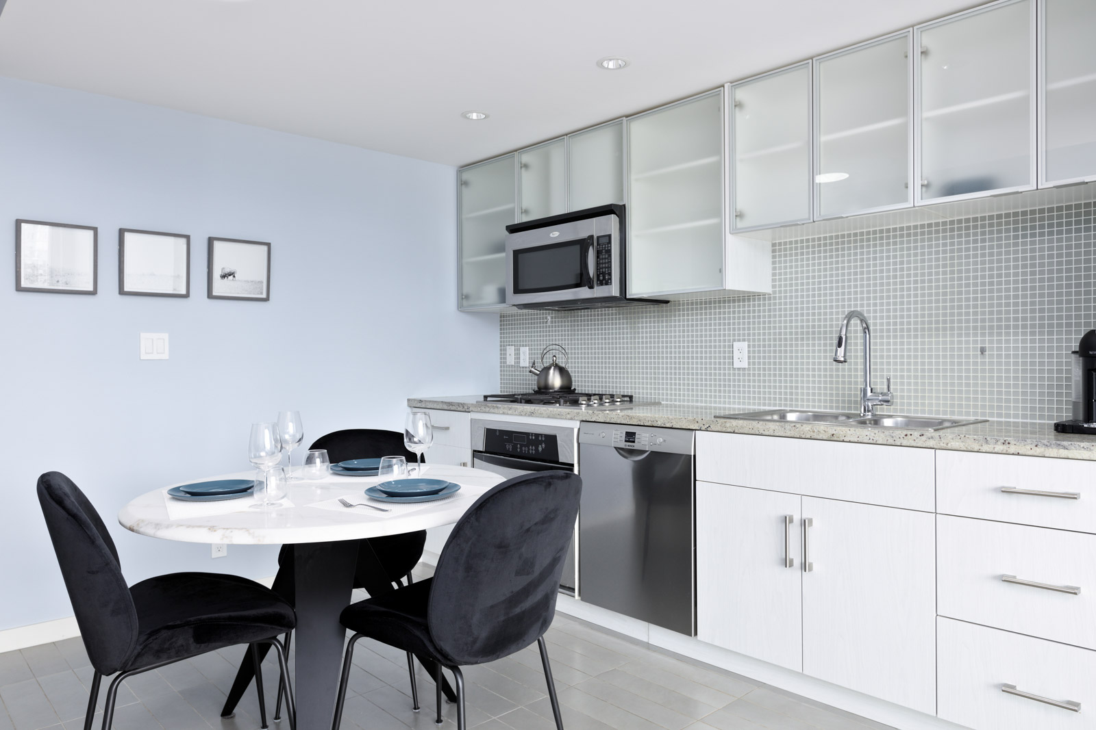 Modern kitchen with light blue walls, a round dining table set with plates and glasses, black chairs, and a kitchenette featuring a stove, dishwasher, microwave, and sink under frosted cabinets.