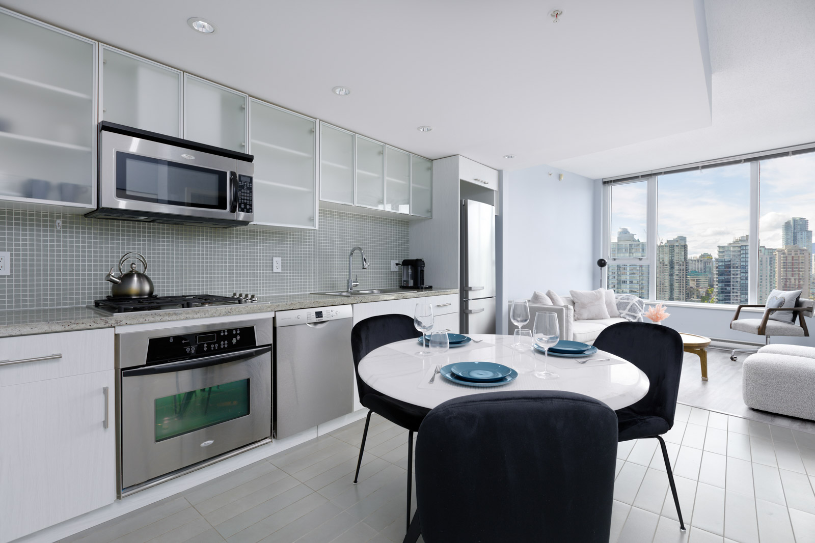 Modern kitchen and dining area with white cabinets, stainless steel appliances, and a round table set for two. Large windows offer a city view in the background.