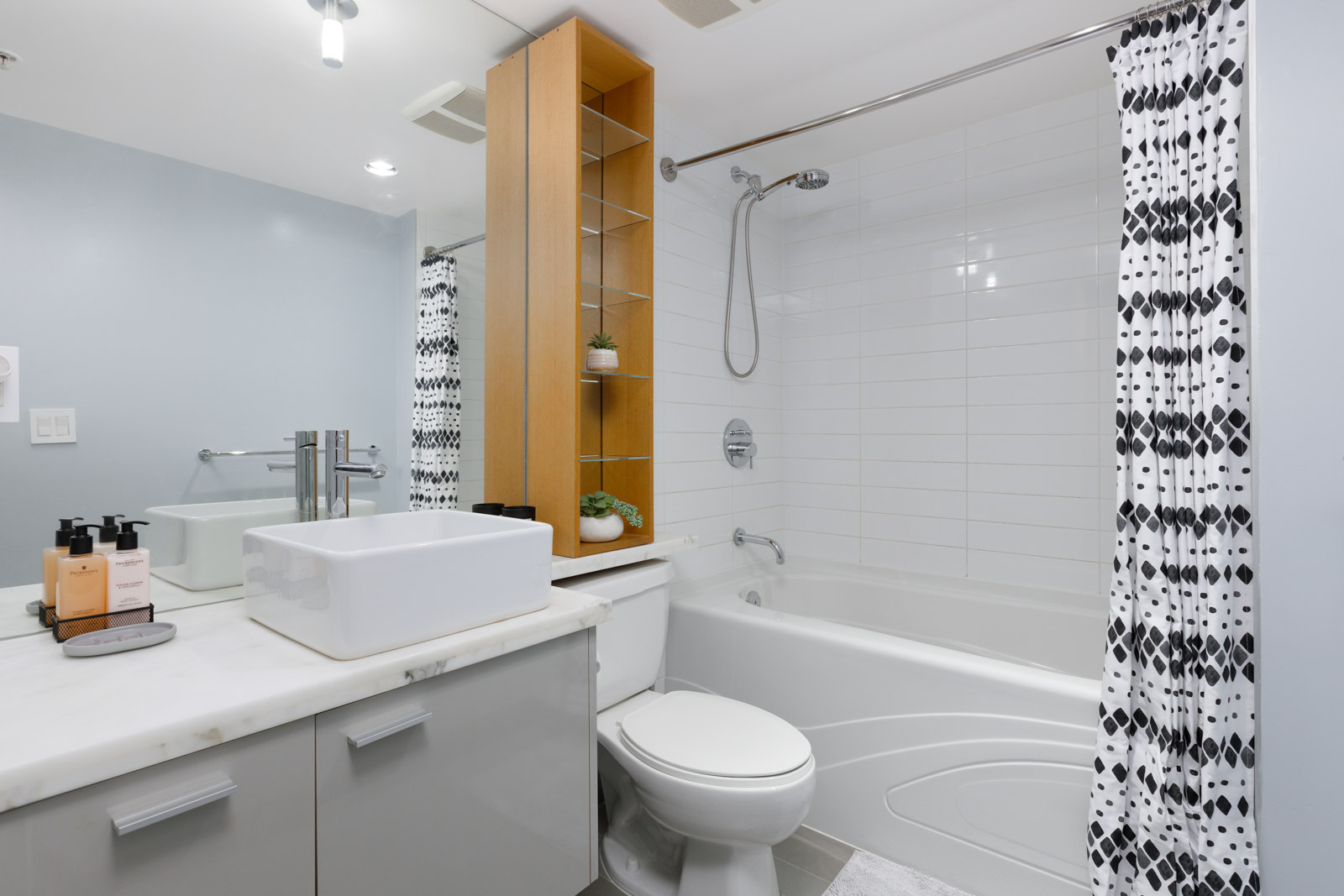 Modern bathroom with wall-mounted sink, white countertop, toilet, and bathtub with geometric-patterned shower curtain. A wooden shelf with plants is above the toilet.