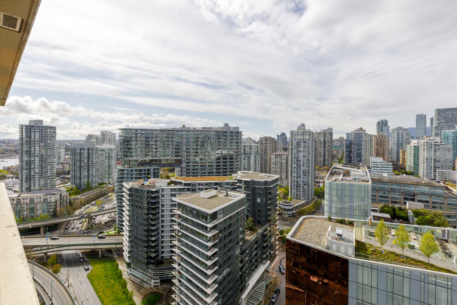 Aerial view of a cityscape featuring modern high-rise buildings, roads, and greenery. Overcast sky with scattered clouds visible above the urban landscape.