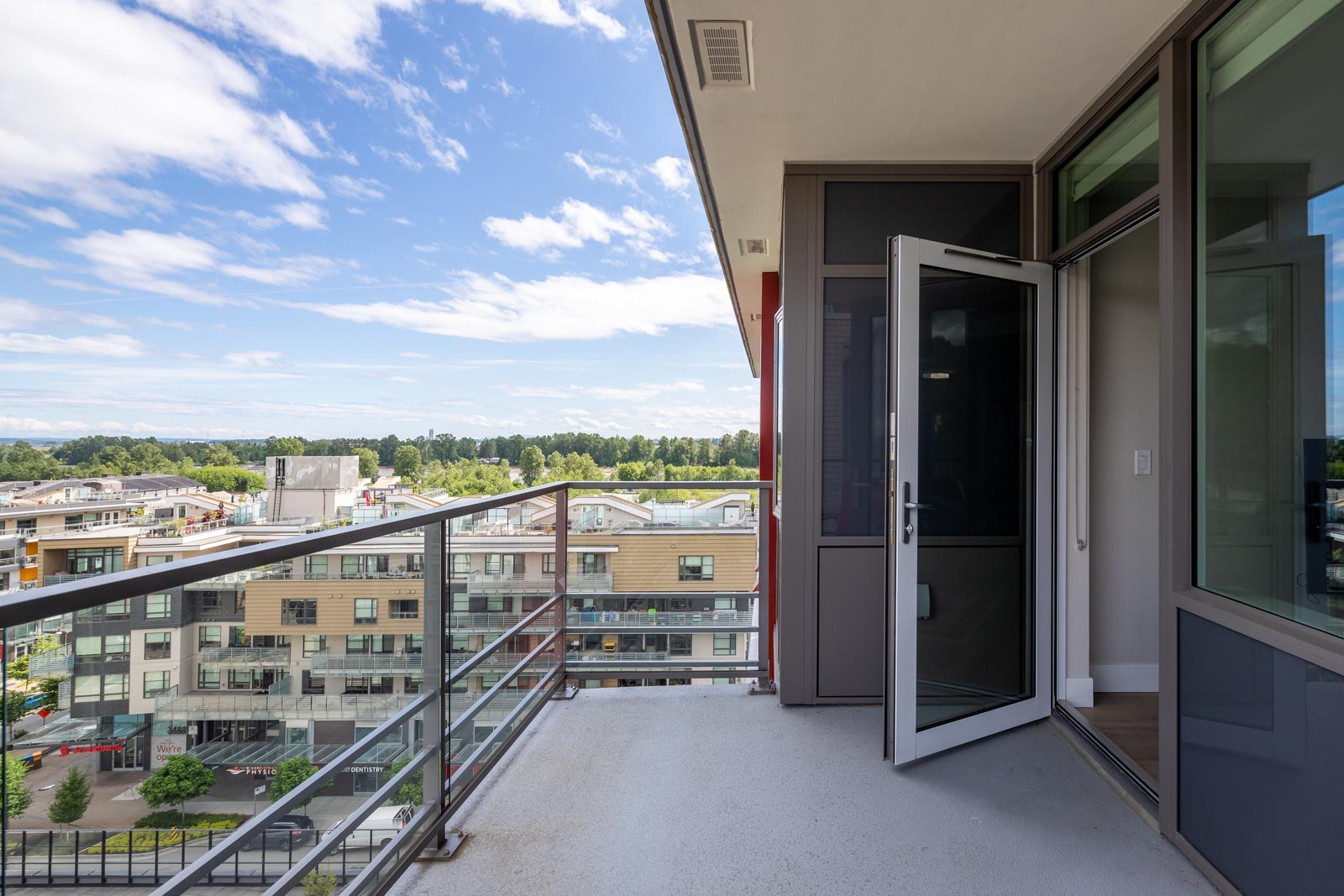 A balcony with metal railings overlooks modern apartment buildings and trees under a partly cloudy sky; a glass door is open to the interior.