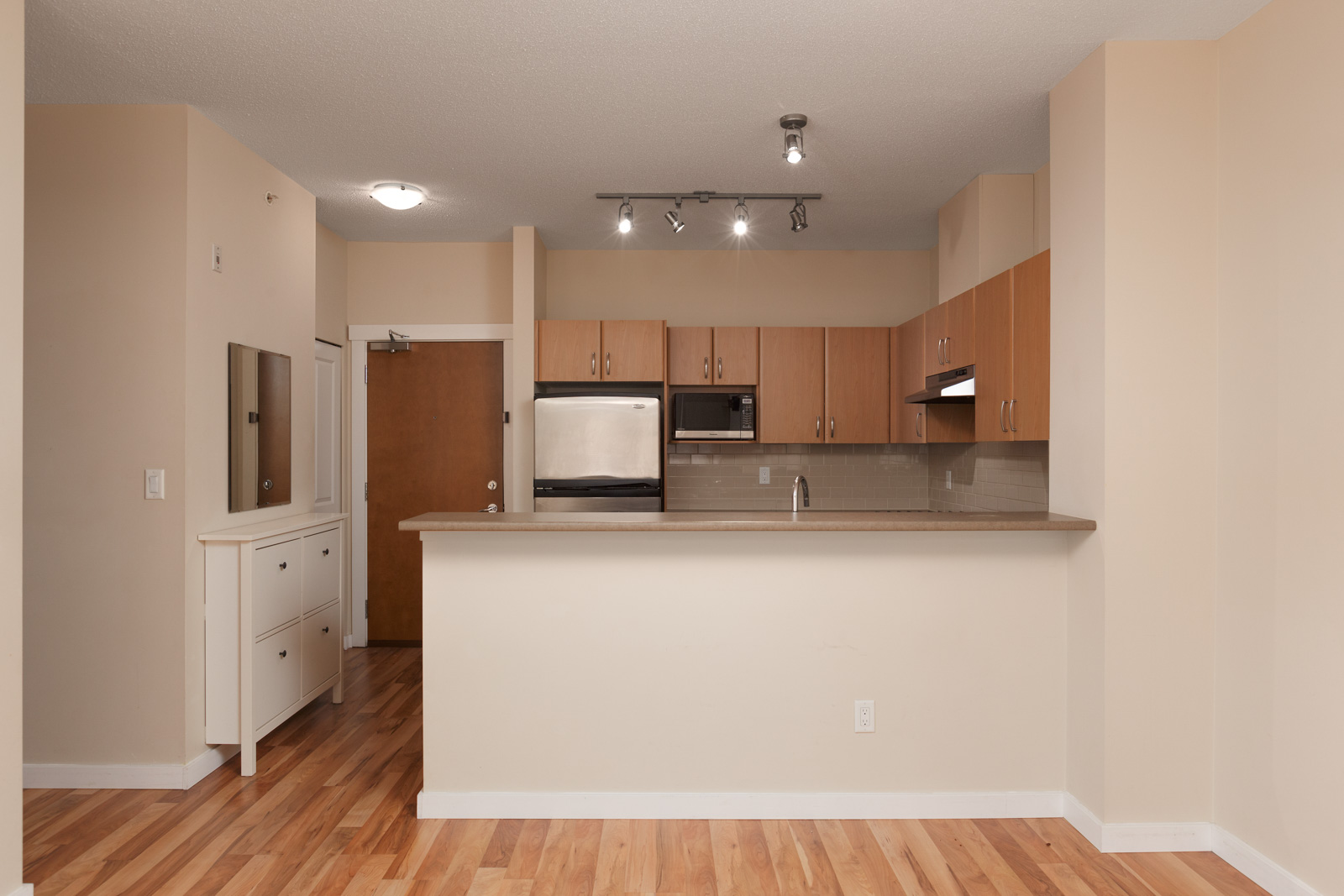 A modern kitchen with wooden cabinets, stainless steel appliances, and a gray countertop. The room features beige walls and hardwood floors.