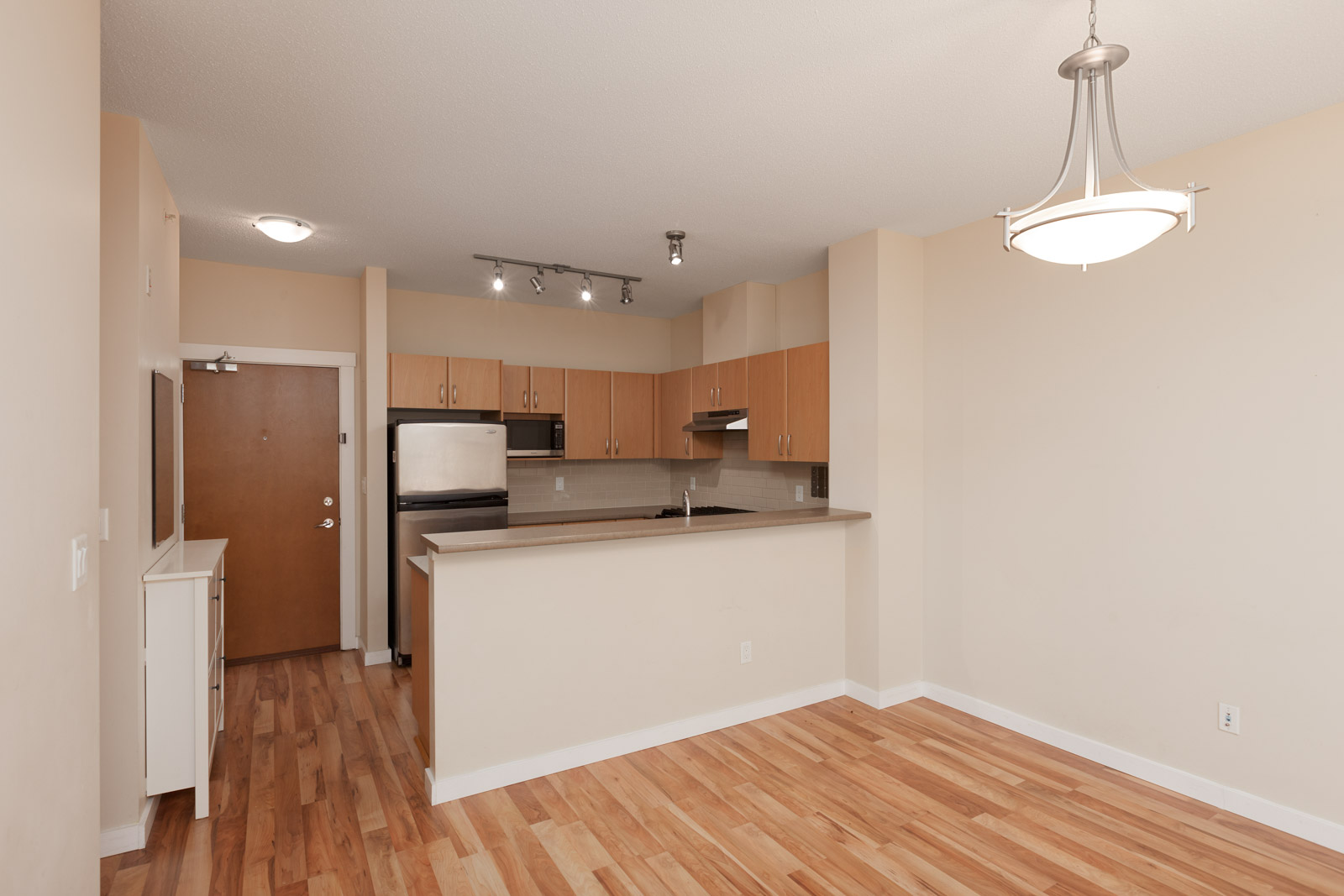 Empty kitchen and dining area with hardwood floors, wooden cabinets, stainless steel appliances, and a light fixture above.