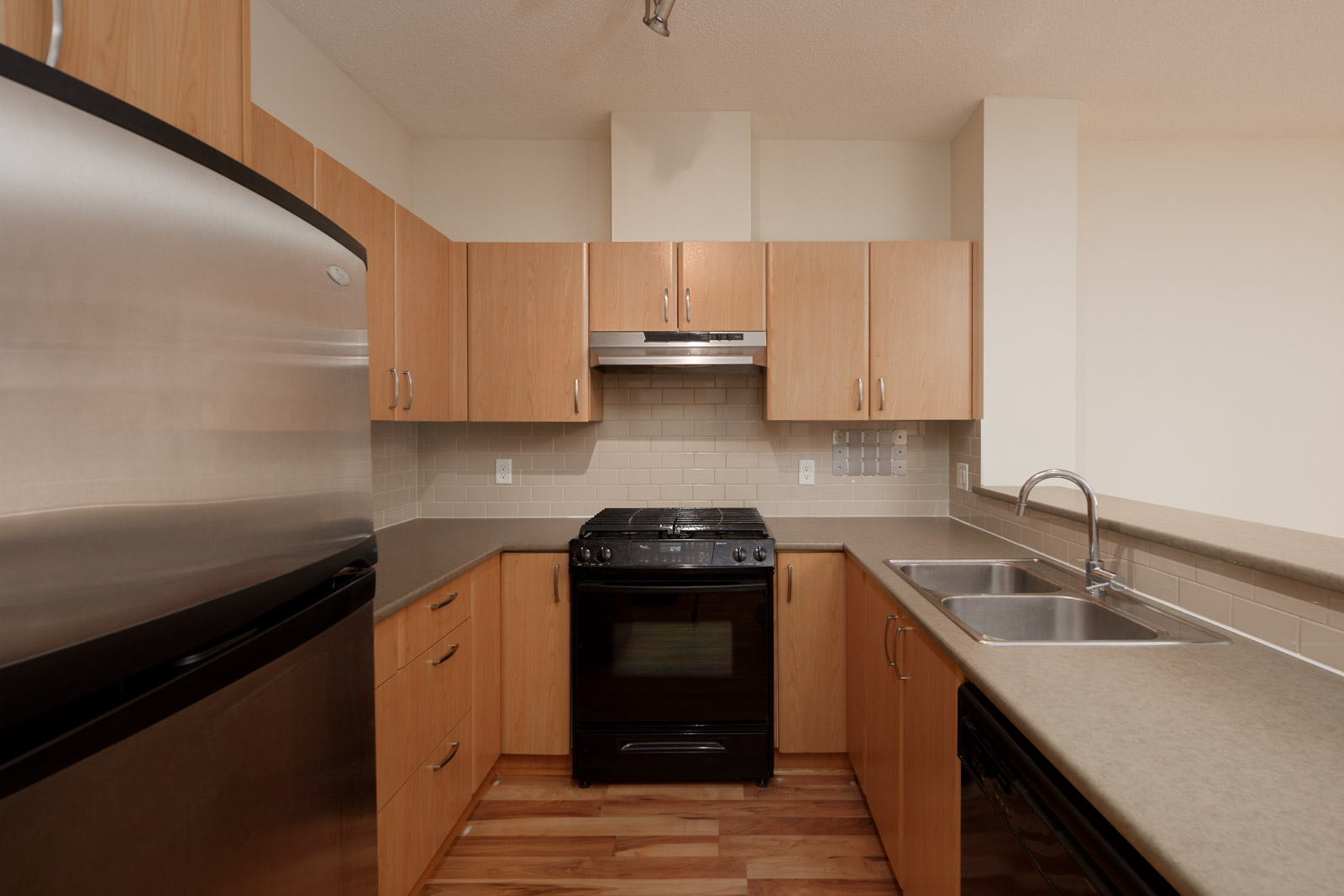 A modern kitchen with wooden cabinets, stainless steel refrigerator, black oven, dual sink, and a beige countertop.