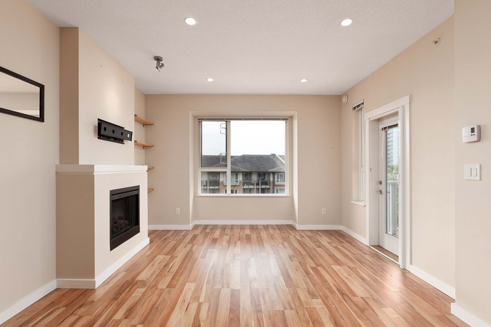 Empty living room with hardwood floors, beige walls, a fireplace, shelves, large window, and a door leading to a balcony.