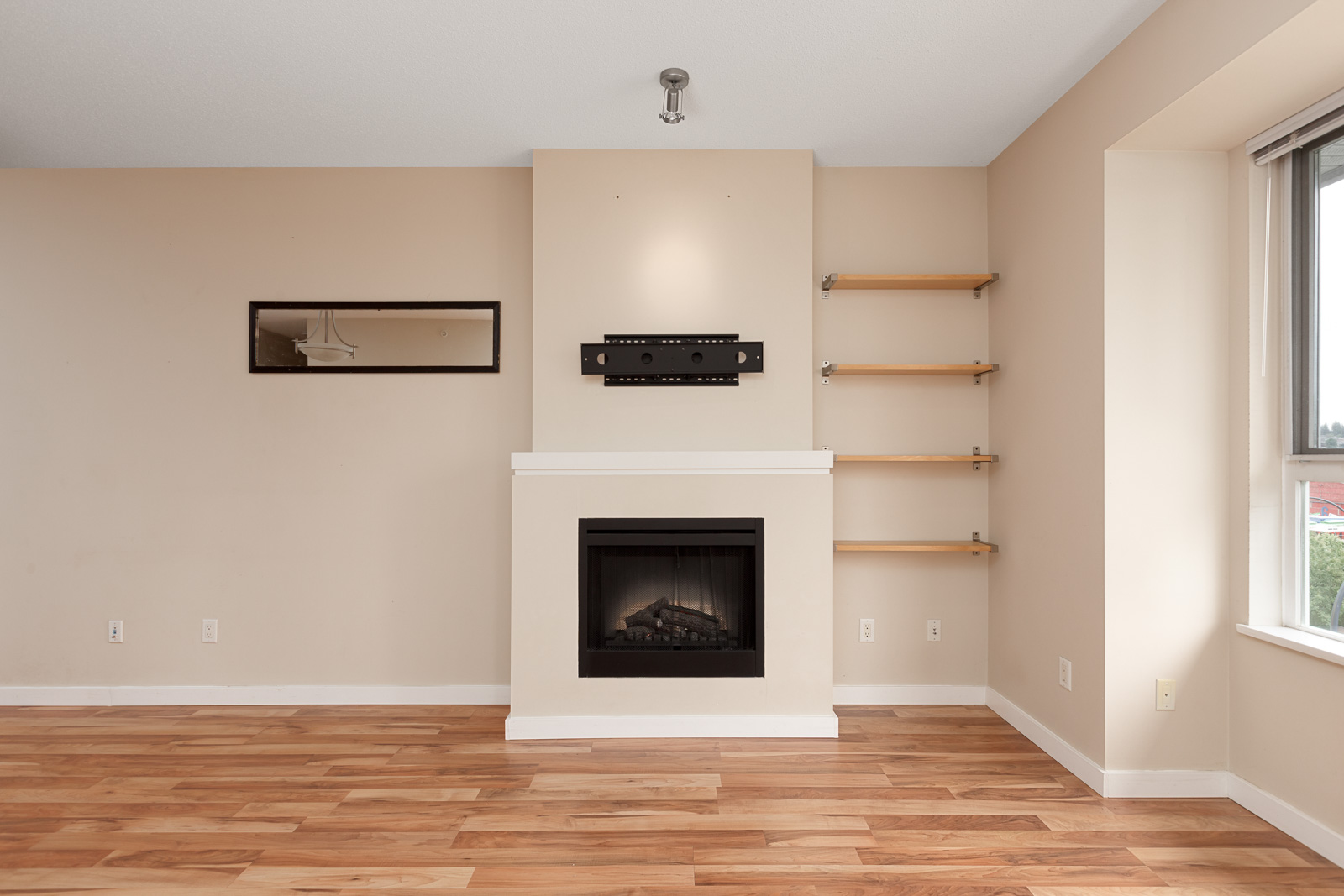 Empty living room with a wall-mounted TV bracket above a fireplace, wooden floor, and shelves to the right. Large window on the right wall.