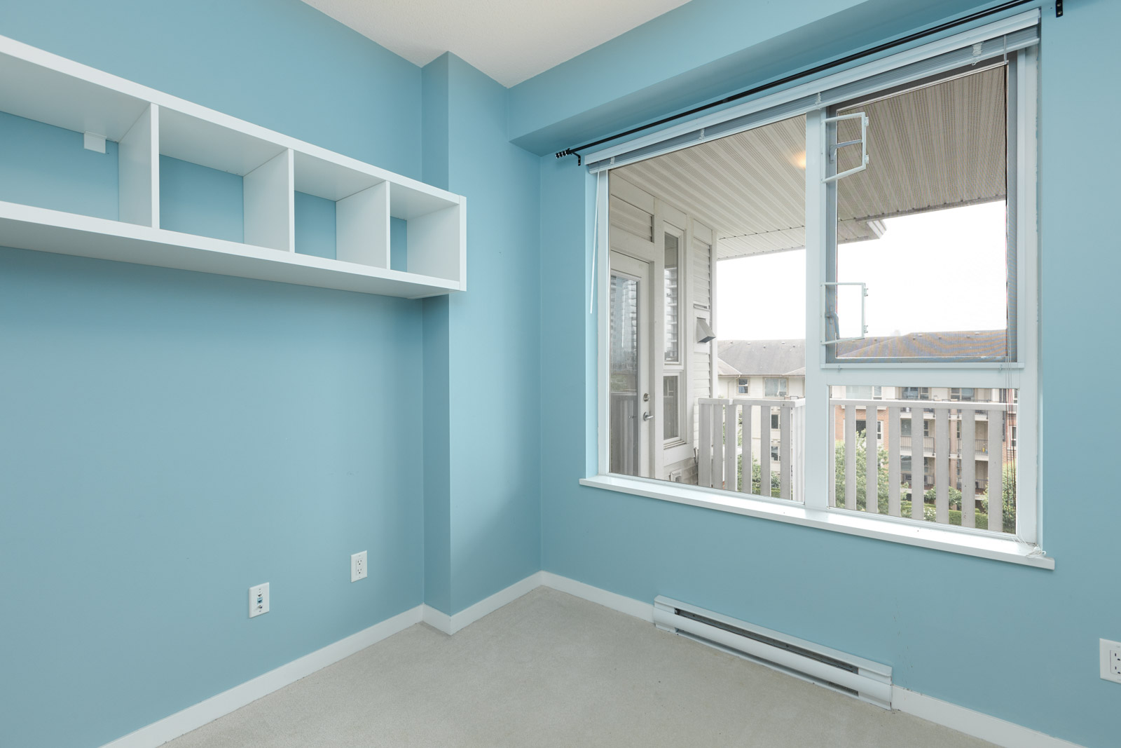 A small, empty room with light blue walls, a shelf, and a large window showing an outdoor view.