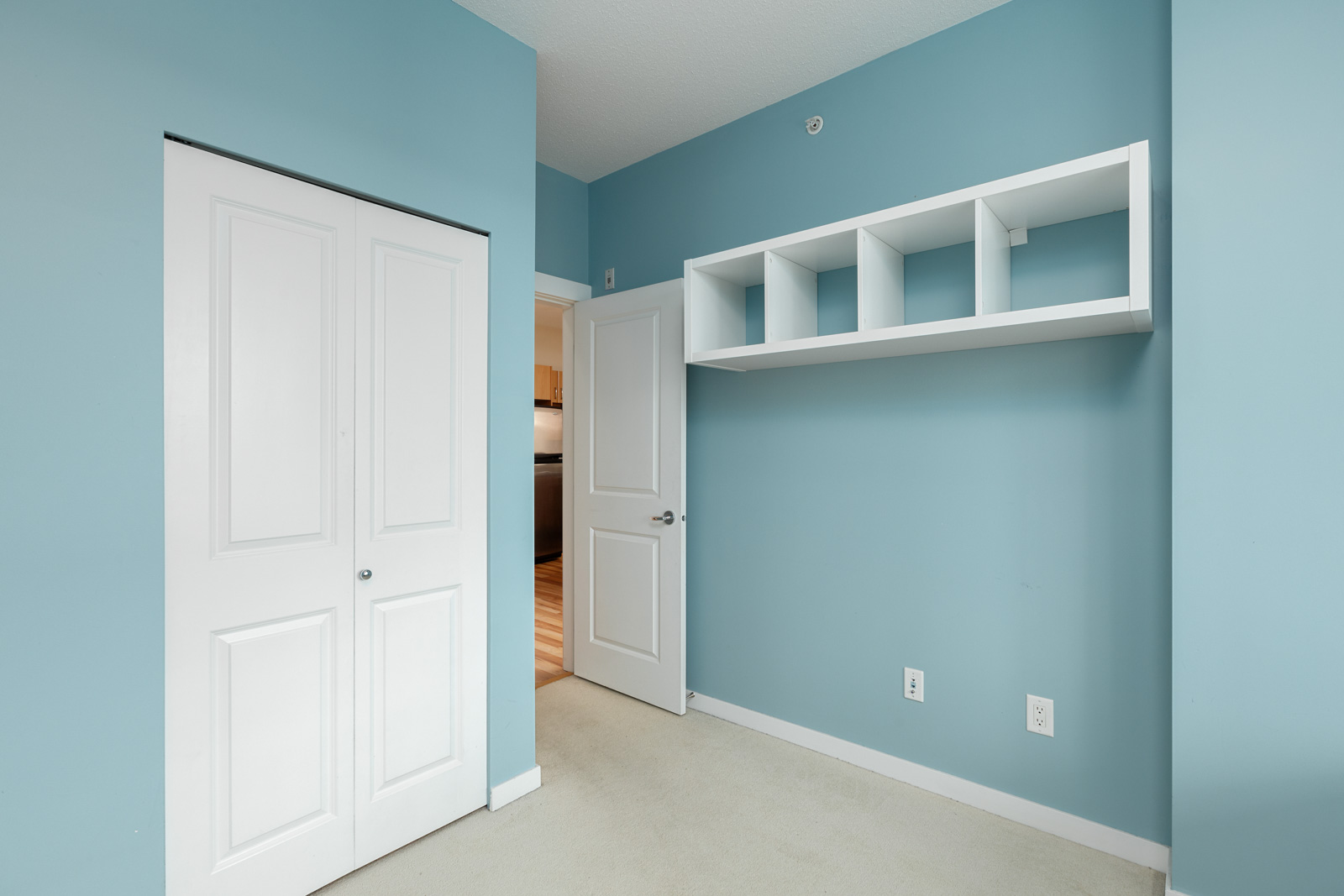 Empty room with light blue walls, white closet doors, and a white shelf. Doorway leads to a wooden floor area.