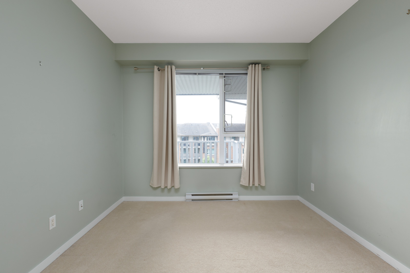 Empty room with pale green walls, beige carpet, and a large window with beige curtains. A building is visible outside.