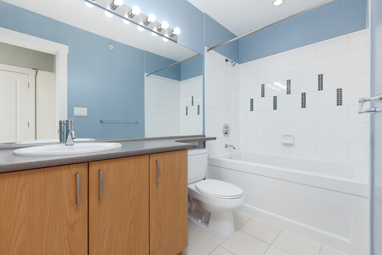 Modern bathroom with blue walls, wood cabinets, white tiles, a white bathtub, and a sink with a large mirror above it.
