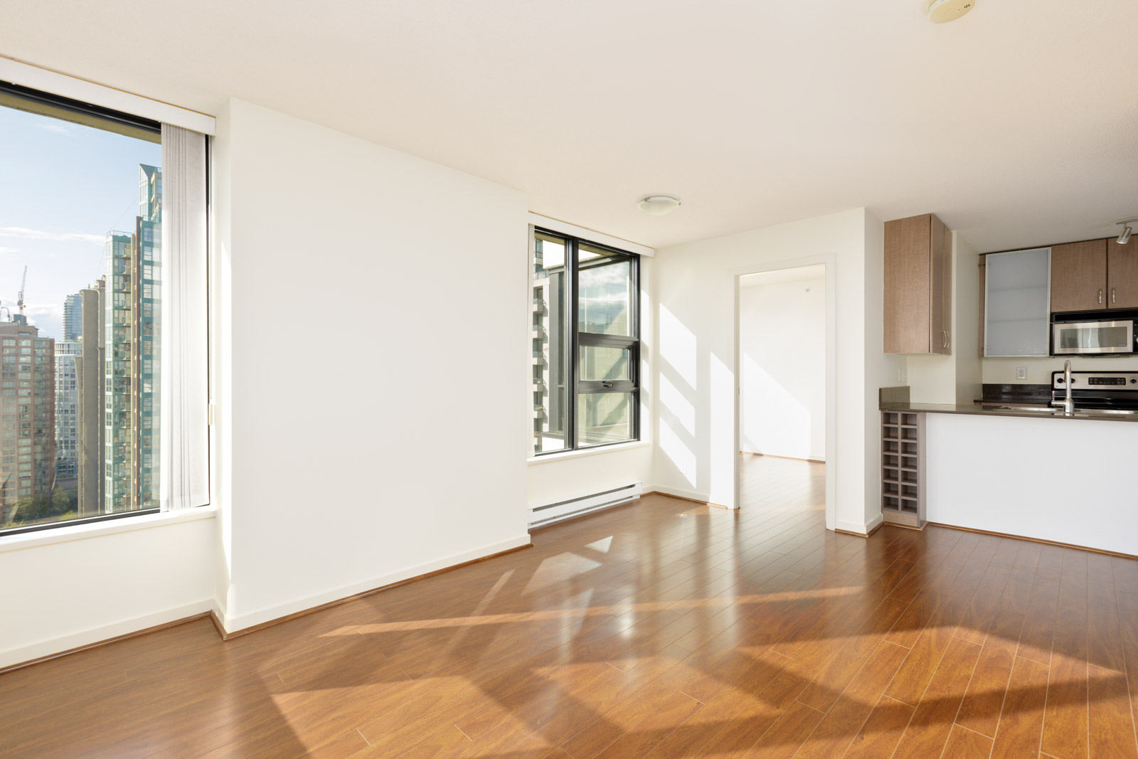 Bright, empty apartment with large windows, wood flooring, white walls, and a modern kitchen with stainless steel appliances. City buildings visible through the windows.