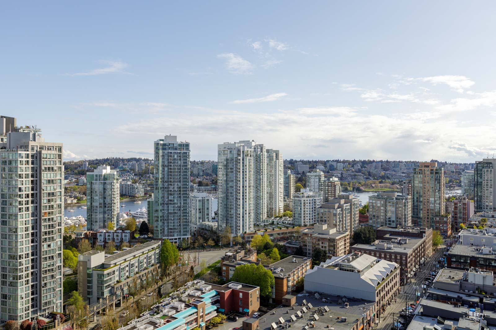 A cityscape with modern high-rise buildings, smaller commercial structures, and a body of water in the background under a partly cloudy sky.