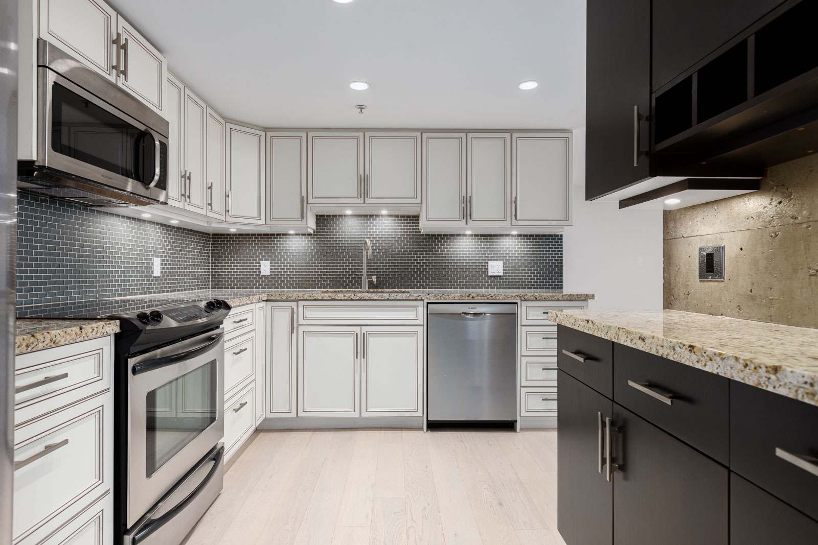 Modern kitchen with white cabinets, stainless steel appliances, granite countertops, light wood flooring, and a gray tile backsplash.