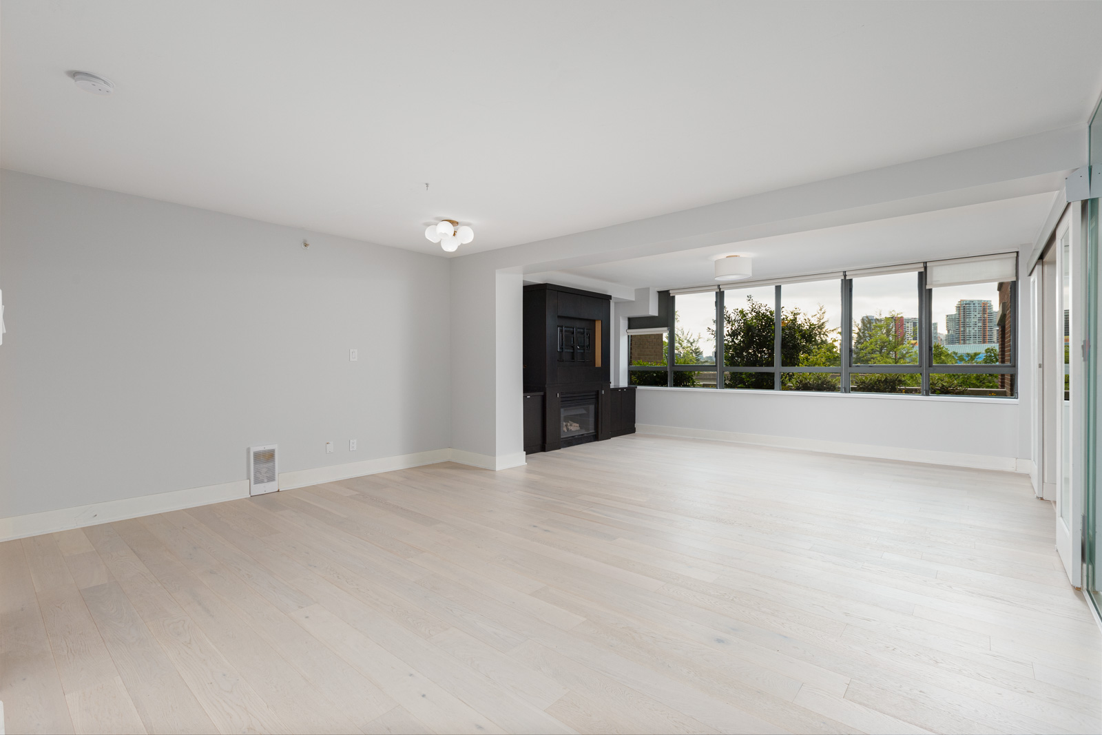 Unfurnished living room with light wood floors, large windows, a black fireplace, and neutral-colored walls, offering a view of greenery and buildings outside.
