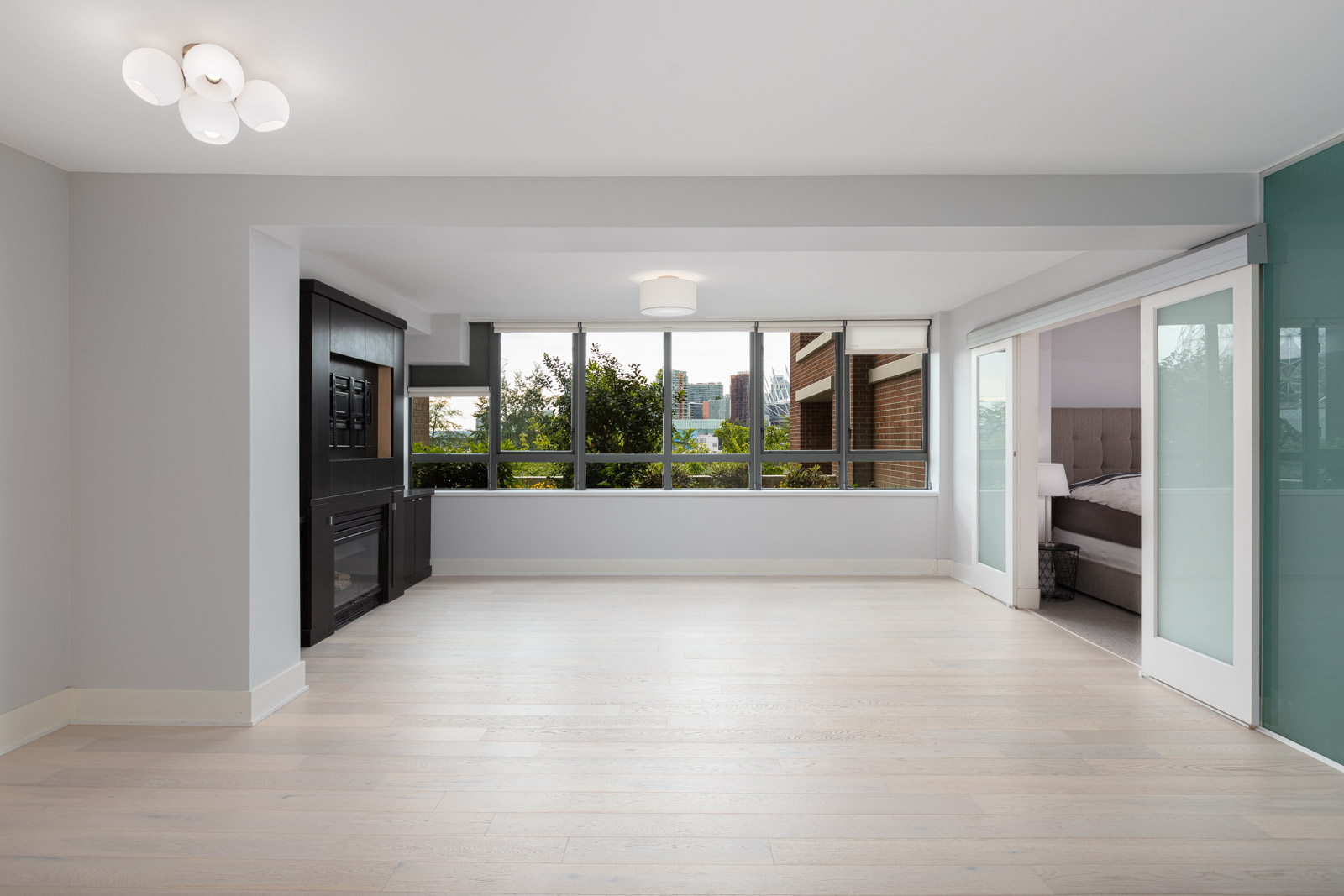 Modern, empty living room with light wood floors, large windows, a black built-in unit, and an adjacent bedroom visible through sliding glass doors.