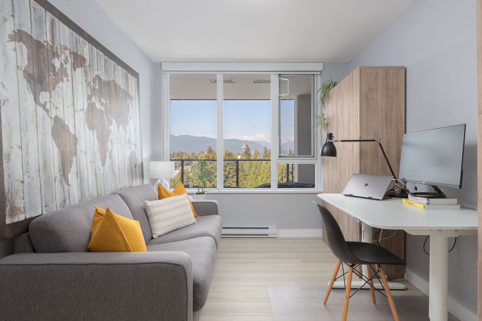 A modern study room with a gray sofa, yellow cushions, a desk with a computer, a desk lamp, and a large world map on the wall. Large windows show a scenic view of mountains and trees.