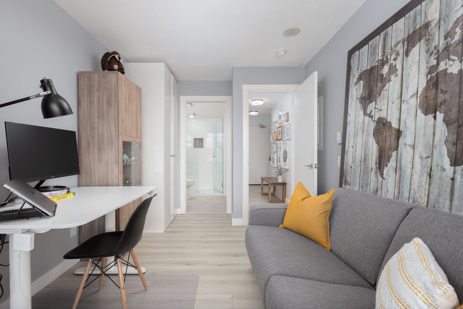 Modern living area with a gray sofa, yellow pillow, desk with computer, and world map wall art. Open door leads to the hallway and bathroom. Light wood flooring throughout.
