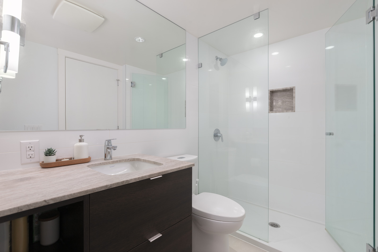 Modern bathroom with glass-enclosed shower, dark wood vanity, white countertop, sink, and a small plant.
