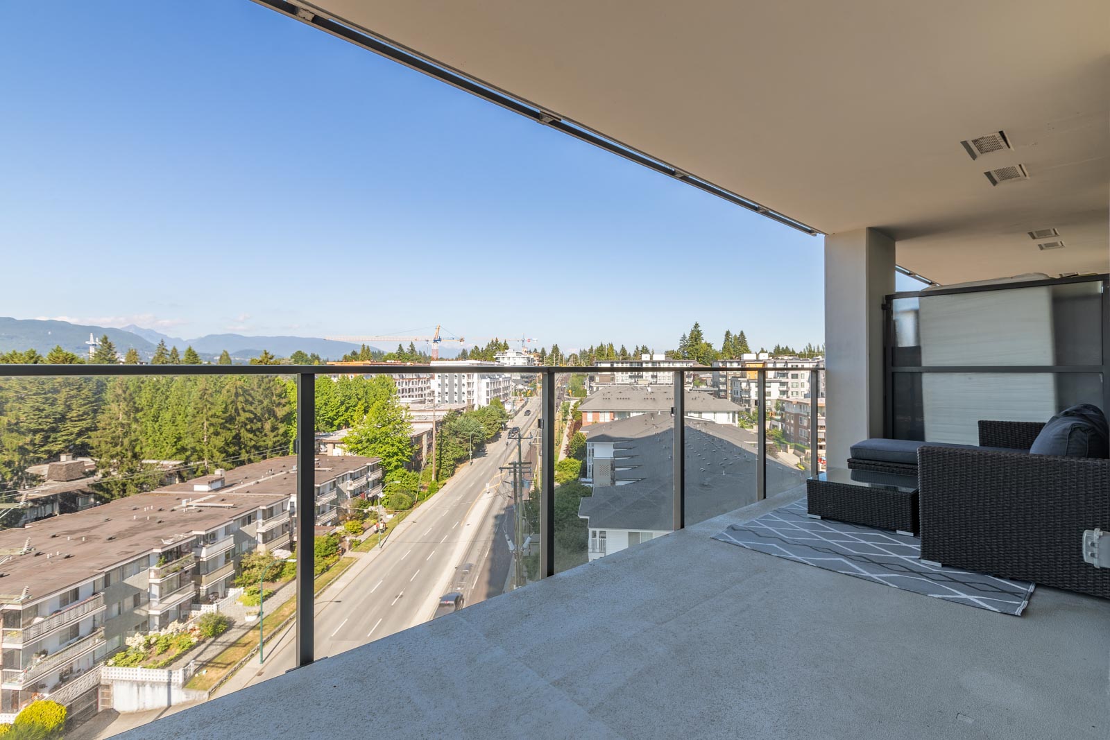 Balcony view overlooking a city street with trees and buildings. Outdoor seating with black couches is on the concrete floor.