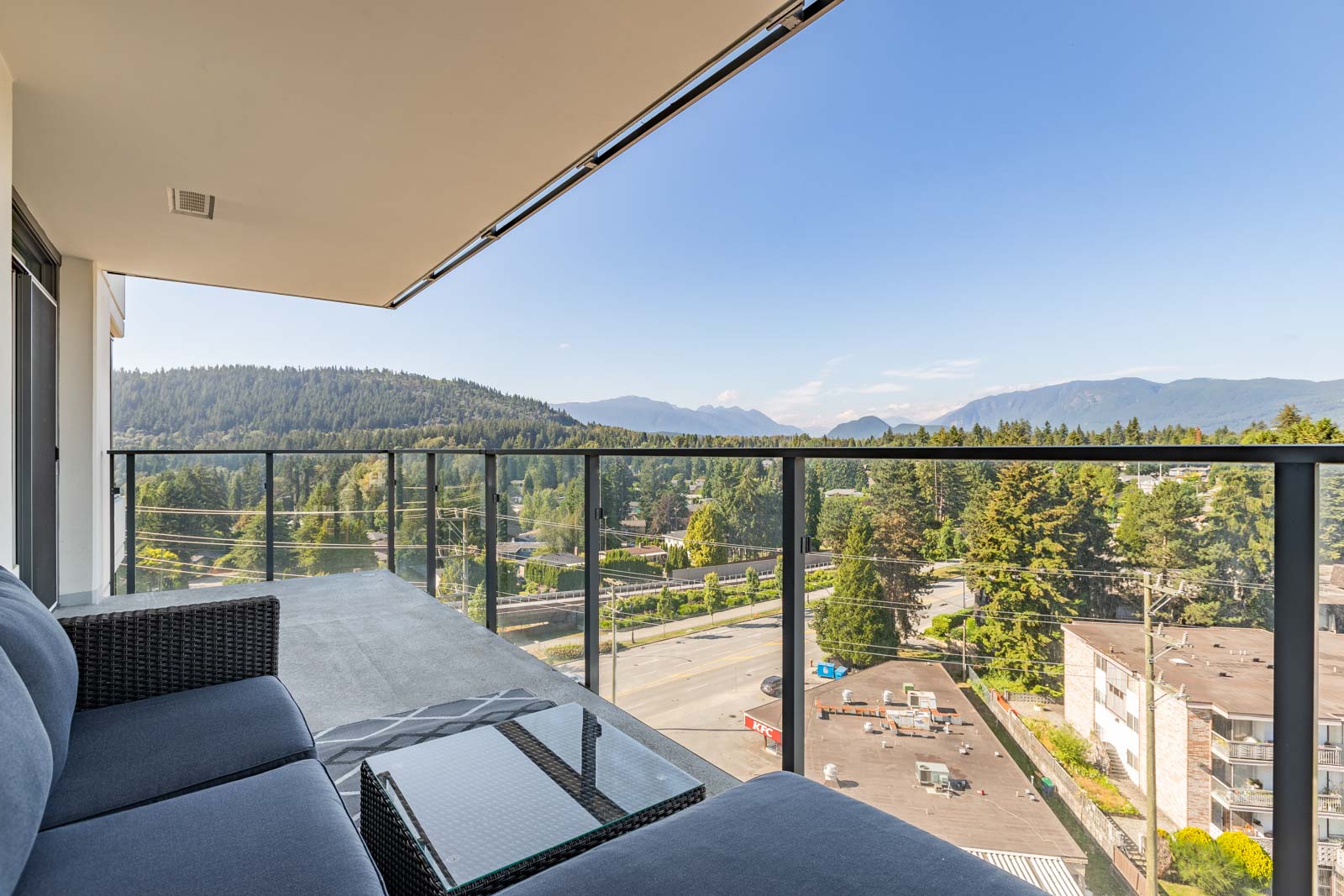 Outdoor balcony with a black wicker couch and glass railing overlooking a view of trees, roads, and distant mountains under a clear blue sky.