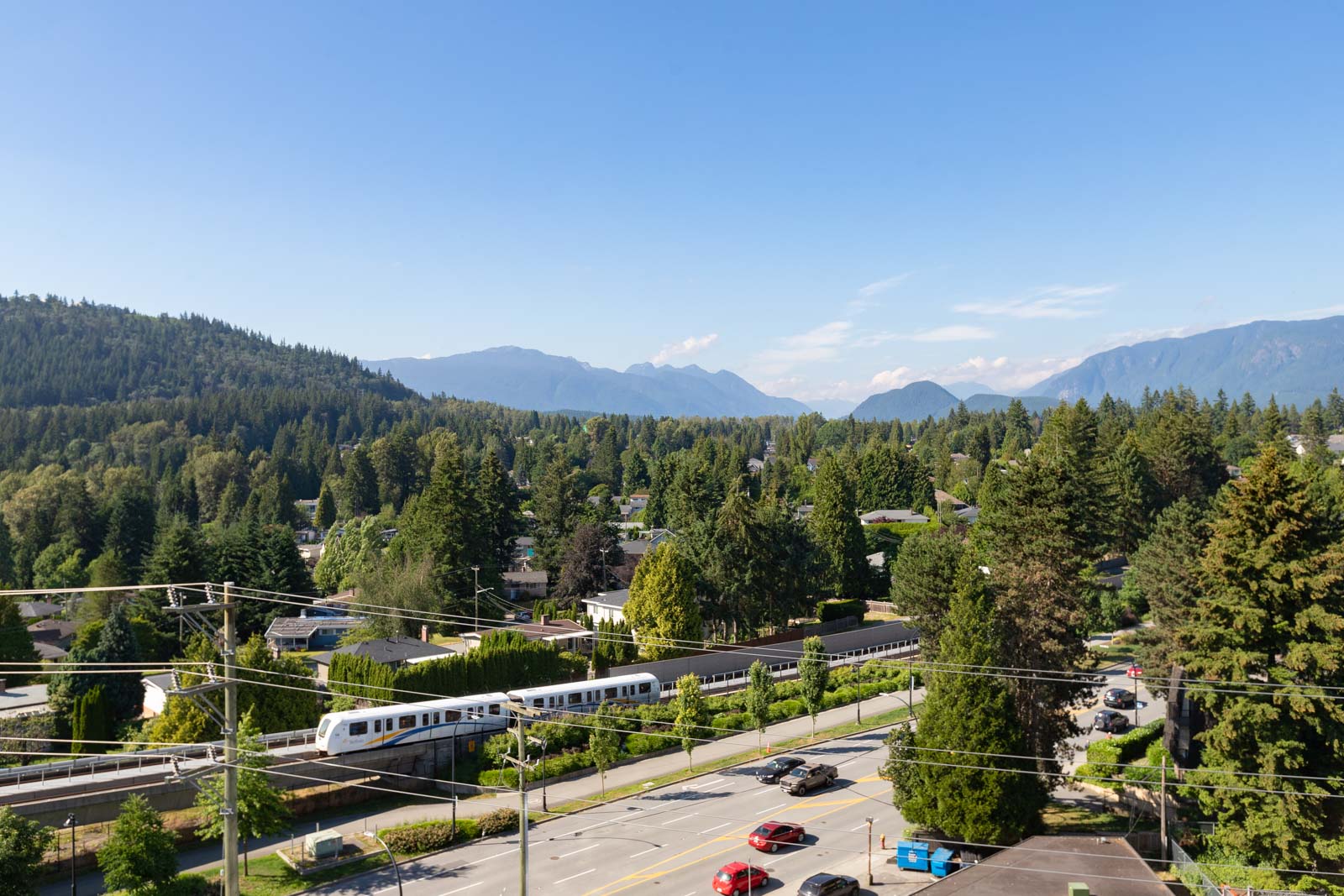 Aerial view of a suburban area with a train passing through, surrounded by trees and mountains under a clear blue sky.