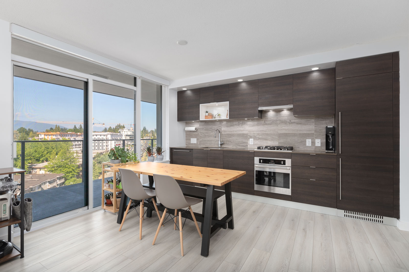 Modern kitchen and dining area with dark wood cabinets, built-in appliances, a wooden table with two chairs, and a large window leading to a balcony with a city view.