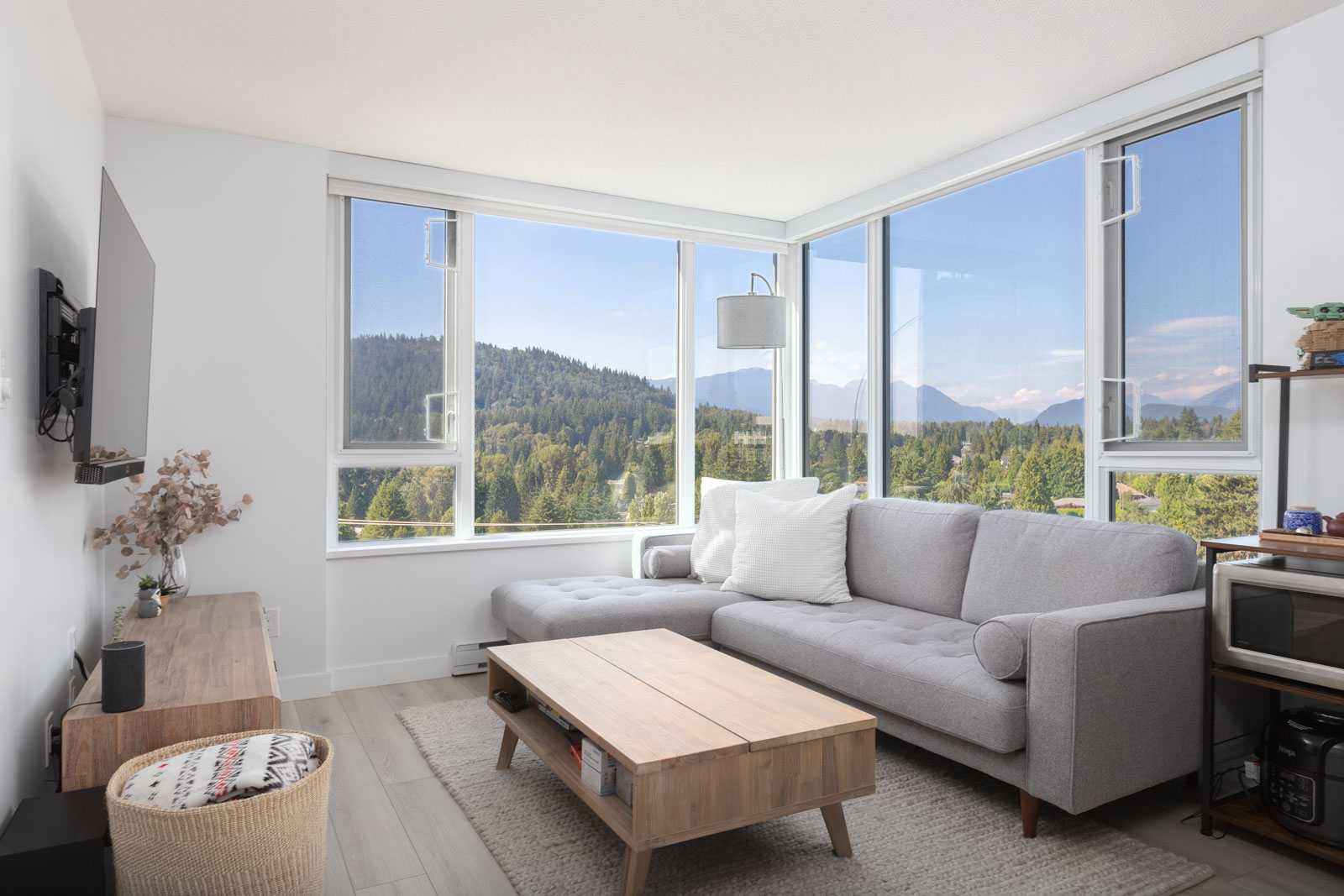 Modern living room with a gray sofa, wooden coffee table, and large windows showcasing a scenic forest and mountain view. A TV is mounted on the wall, and decor items are on a shelf.