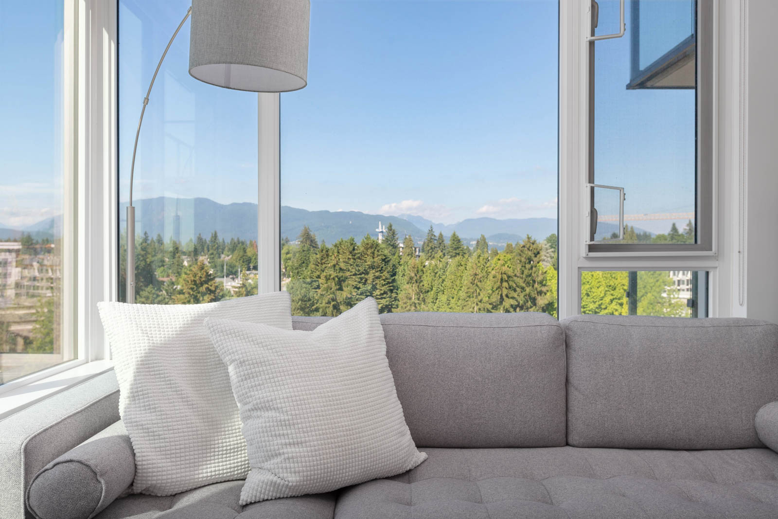 A gray sofa with white pillows next to a floor lamp in a room with large windows, offering a view of trees and distant mountains under a clear blue sky.