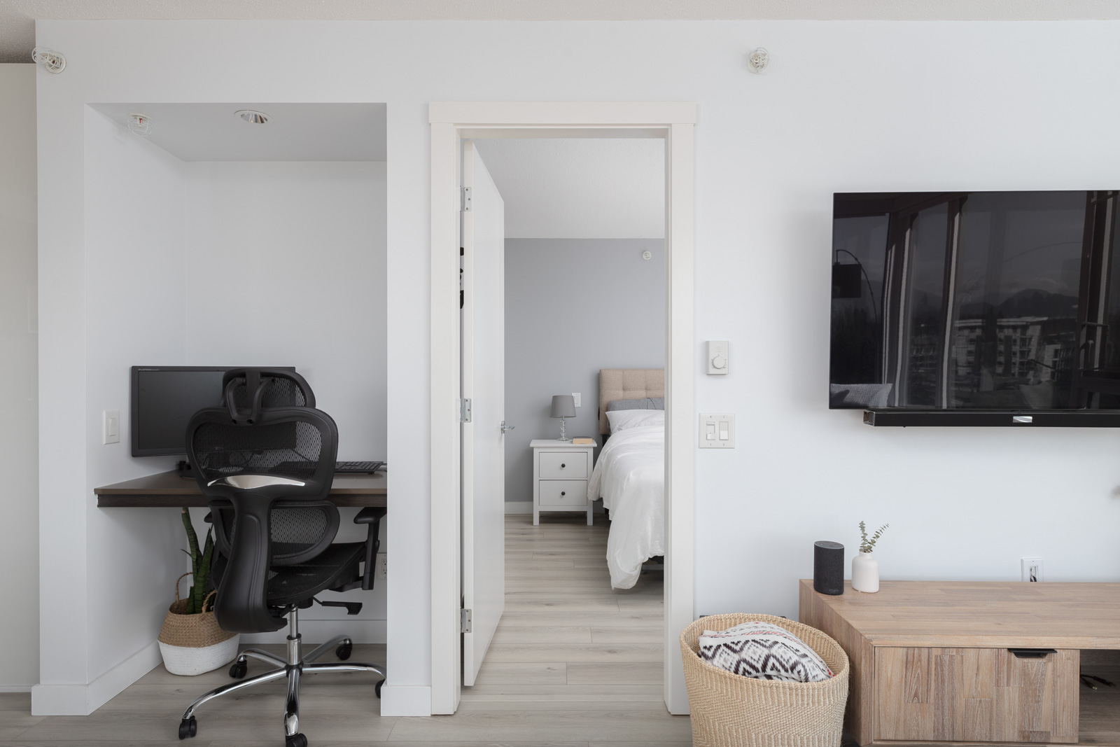 Modern room with a desk and chair in one area and a bed behind an open door. A TV is mounted on the wall, and baskets are on the floor. Neutral color scheme with natural light.