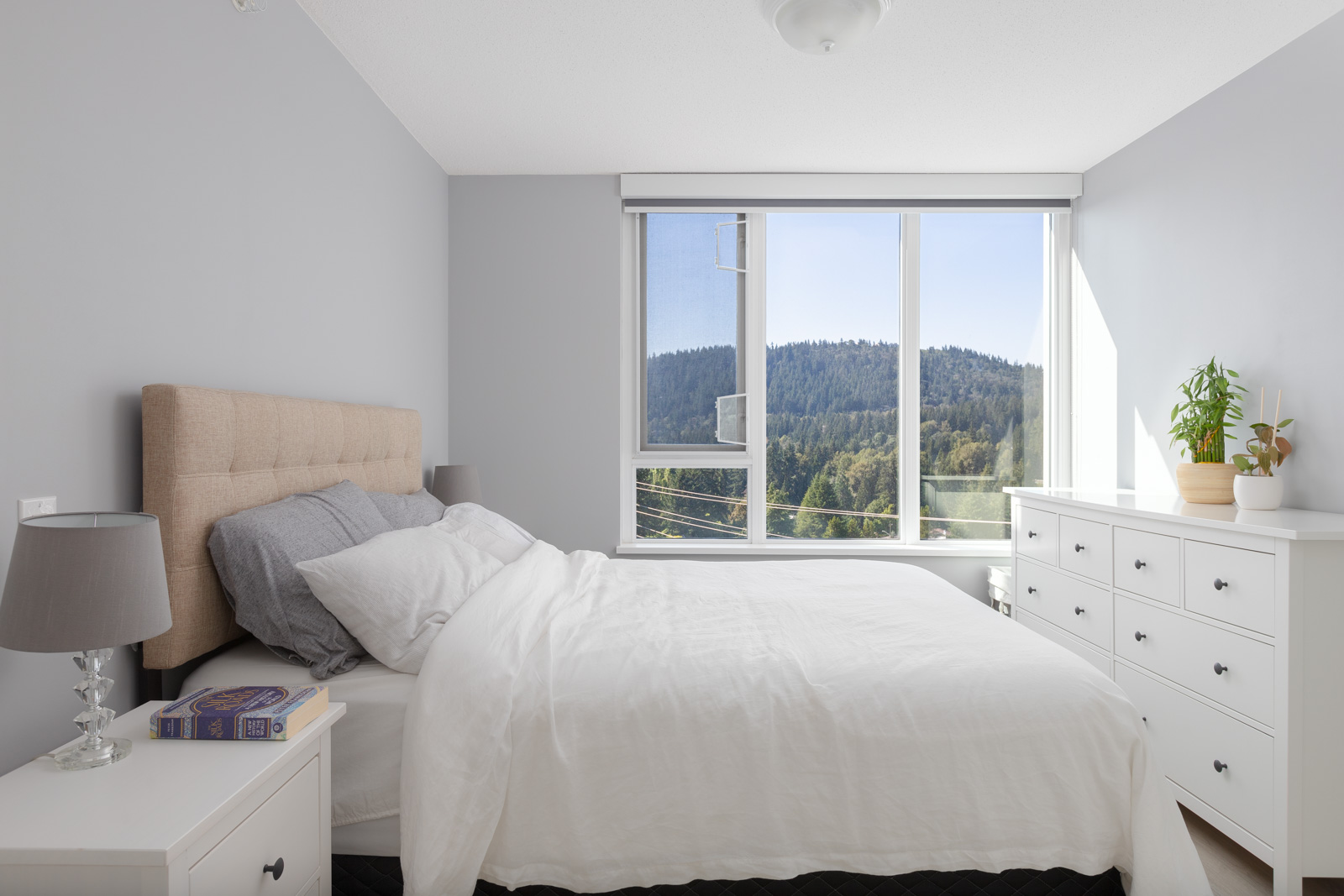 A neatly arranged bedroom with a bed, white linens, nightstand, and dresser. A large window shows a view of forested hills. Lamp and plant decorate the dresser.