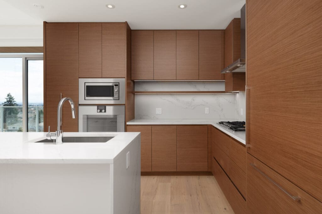 Modern kitchen with wood cabinets, white marble countertops and backsplash, built-in microwave, oven, and a stainless steel sink on a central island. Large window with outdoor view.