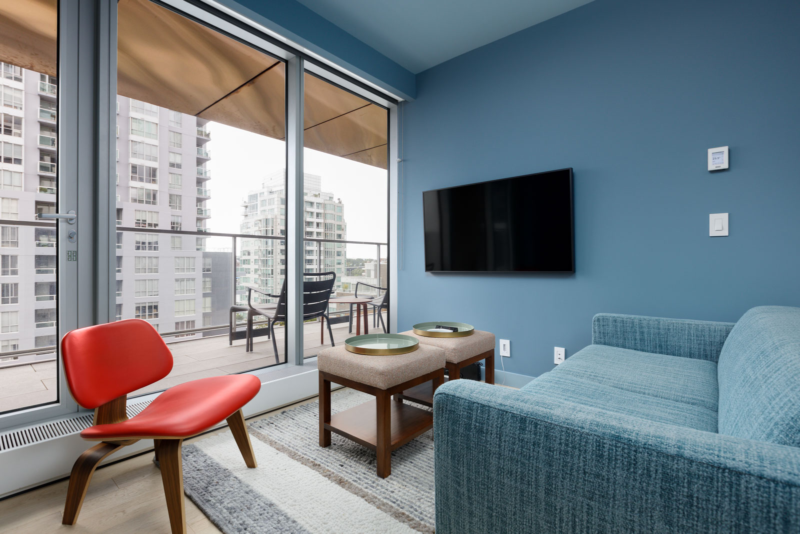 Modern living room with a blue sofa, red chair, and two tables facing a wall-mounted TV. Floor-to-ceiling windows open to a balcony with outdoor seating and city views.
