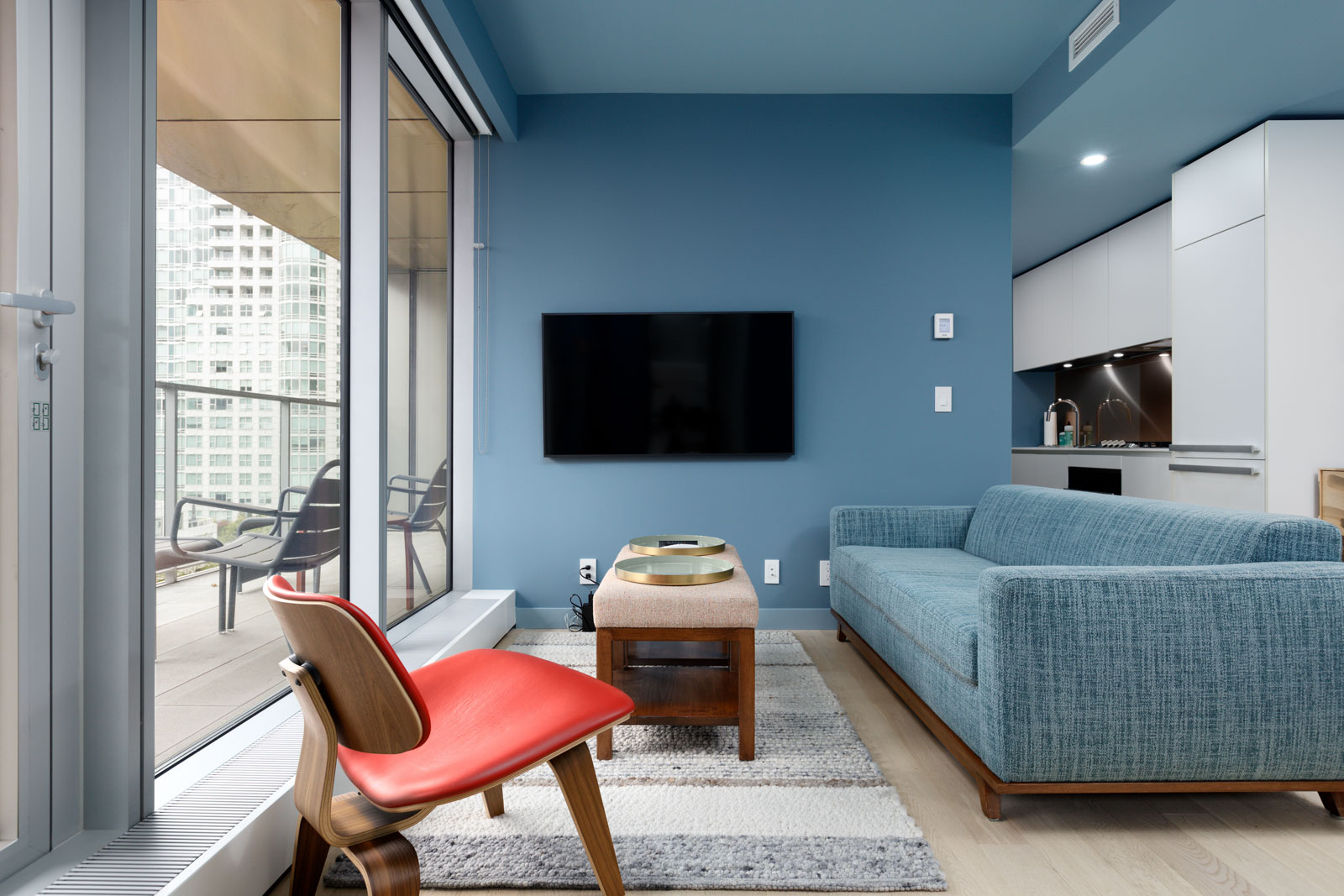 Modern living room with blue walls, a flat-screen TV, blue sofa, wooden coffee table, red chair, and a sliding glass door leading to a balcony with city view.