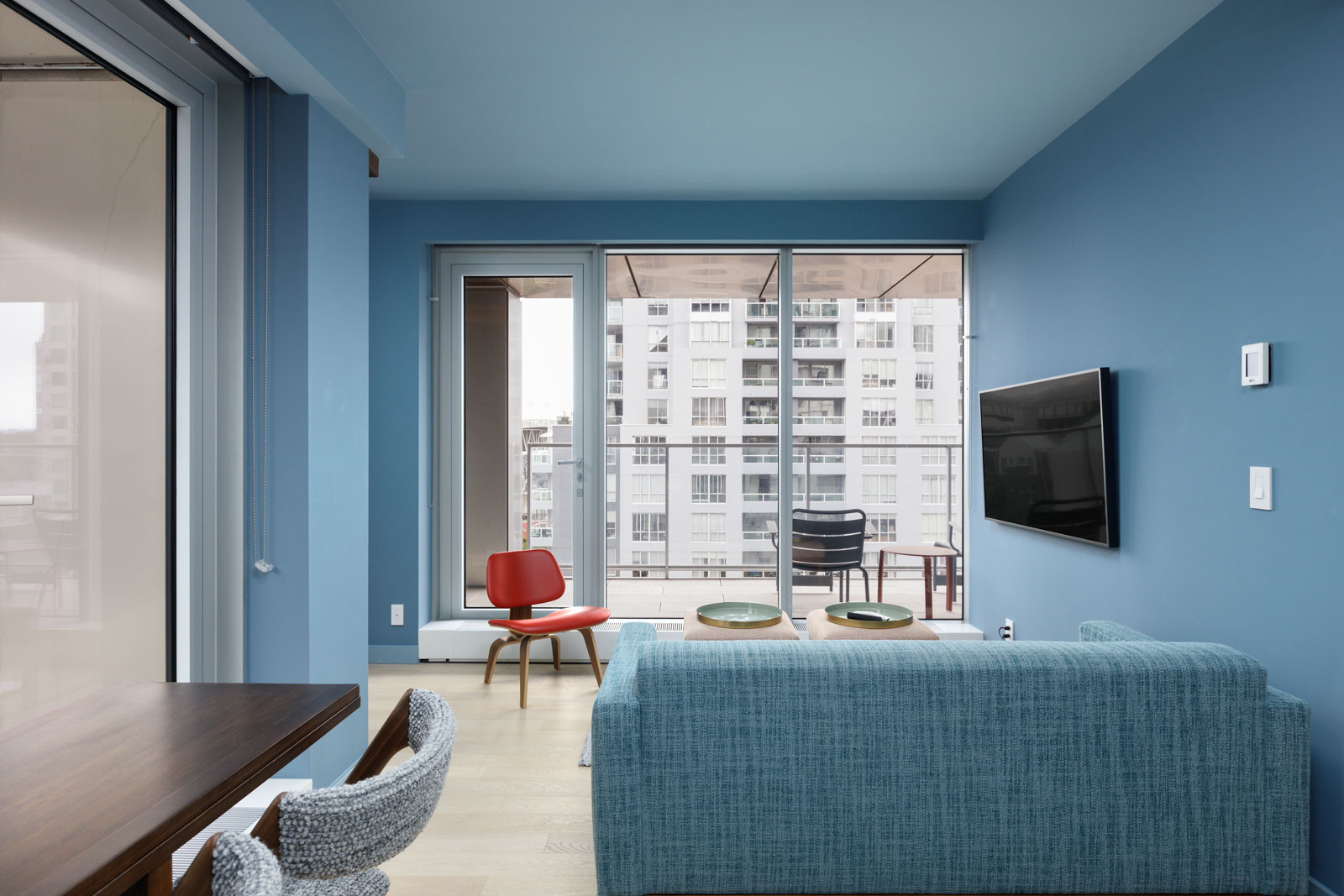 Modern living room with blue walls and sofa, wall-mounted TV, and large windows. A red chair and a small table with two plates are near the windows, overlooking a cityscape.
